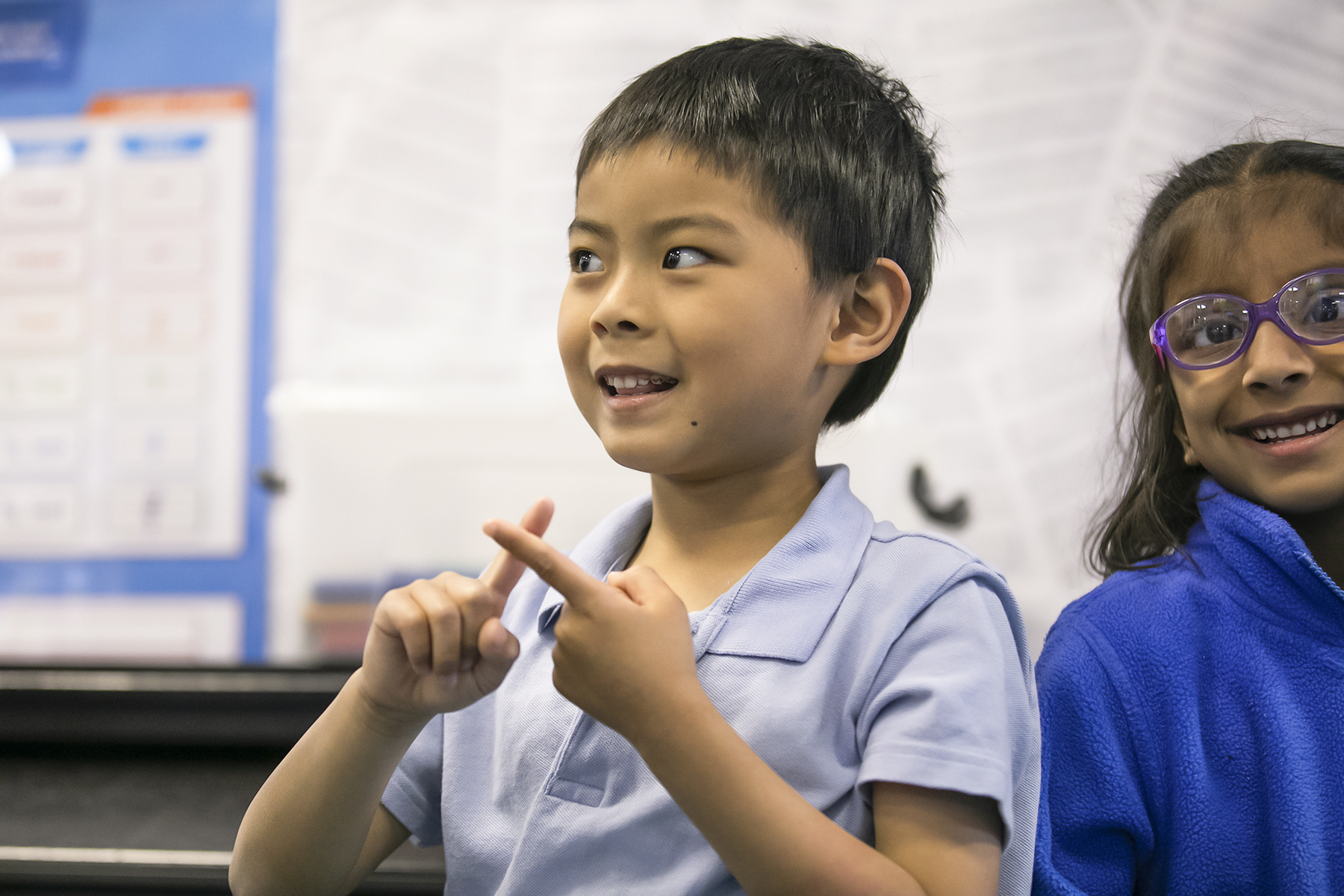 In2Music.GM_.Singlegate.16.COL_.sm_ A young boy in a light blue shirt is using his fingers to count or gesture, while smiling. Beside him, a girl in glasses and a blue fleece smiles as well. They appear to be in a CPD classroom setting.