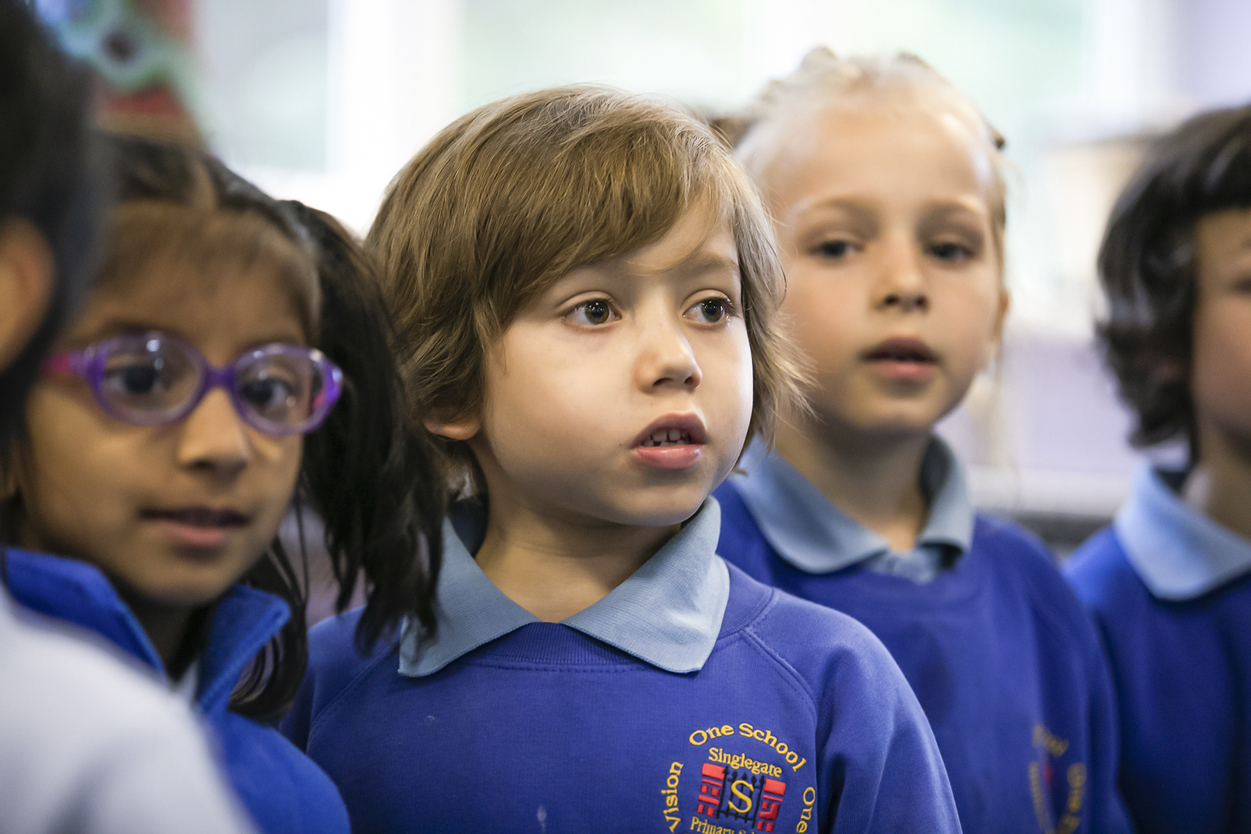 In2Music.GM_.Singlegate.20.COL_.sm_ A group of young children in blue school uniforms stands together indoors, looking attentive during a CPD-related activity. One child with light brown hair is in focus, while others appear slightly blurred in the background.