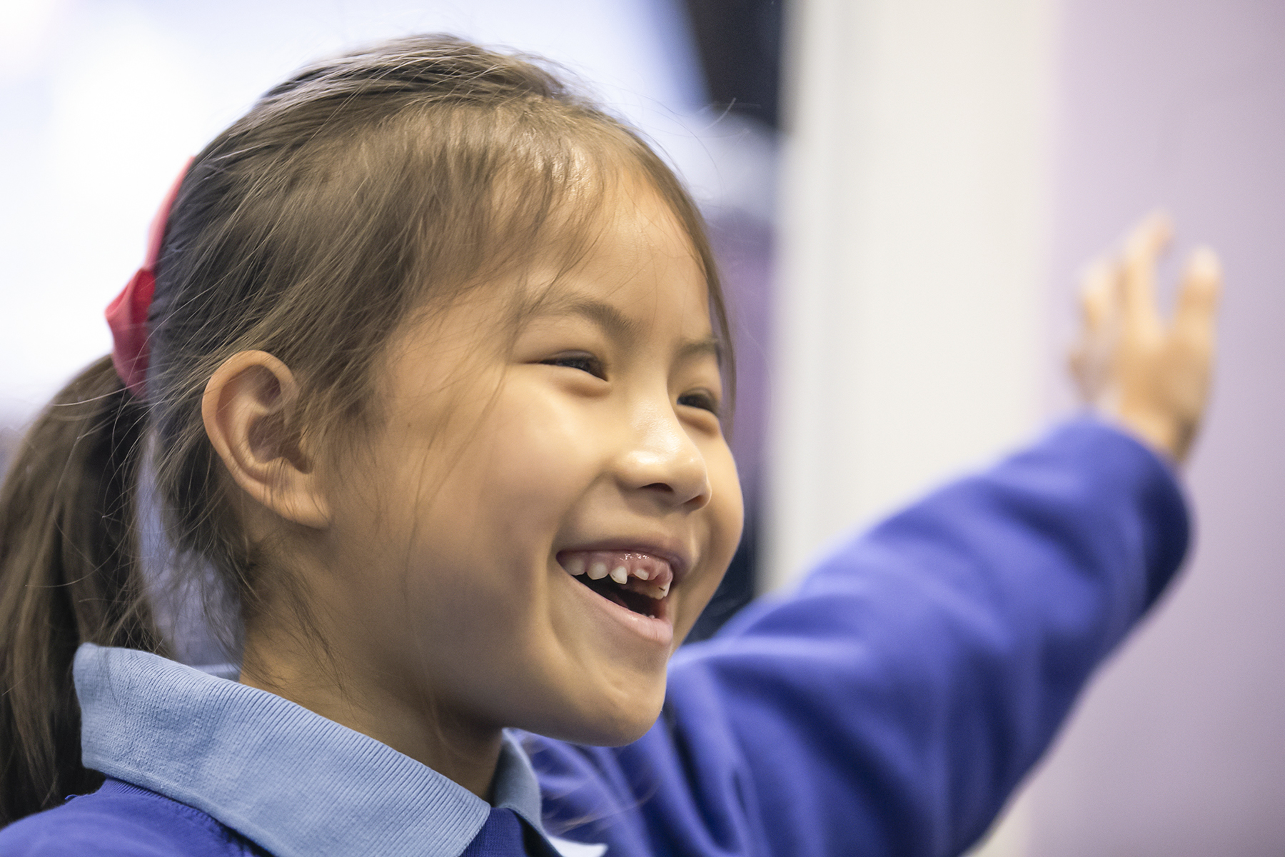 In2Music.GM_.Singlegate.22.COL_.sm_ A young girl with a ponytail and red hair tie smiles brightly and raises her arm, appearing happy and engaged during a CPD activity, while wearing a blue collared shirt.