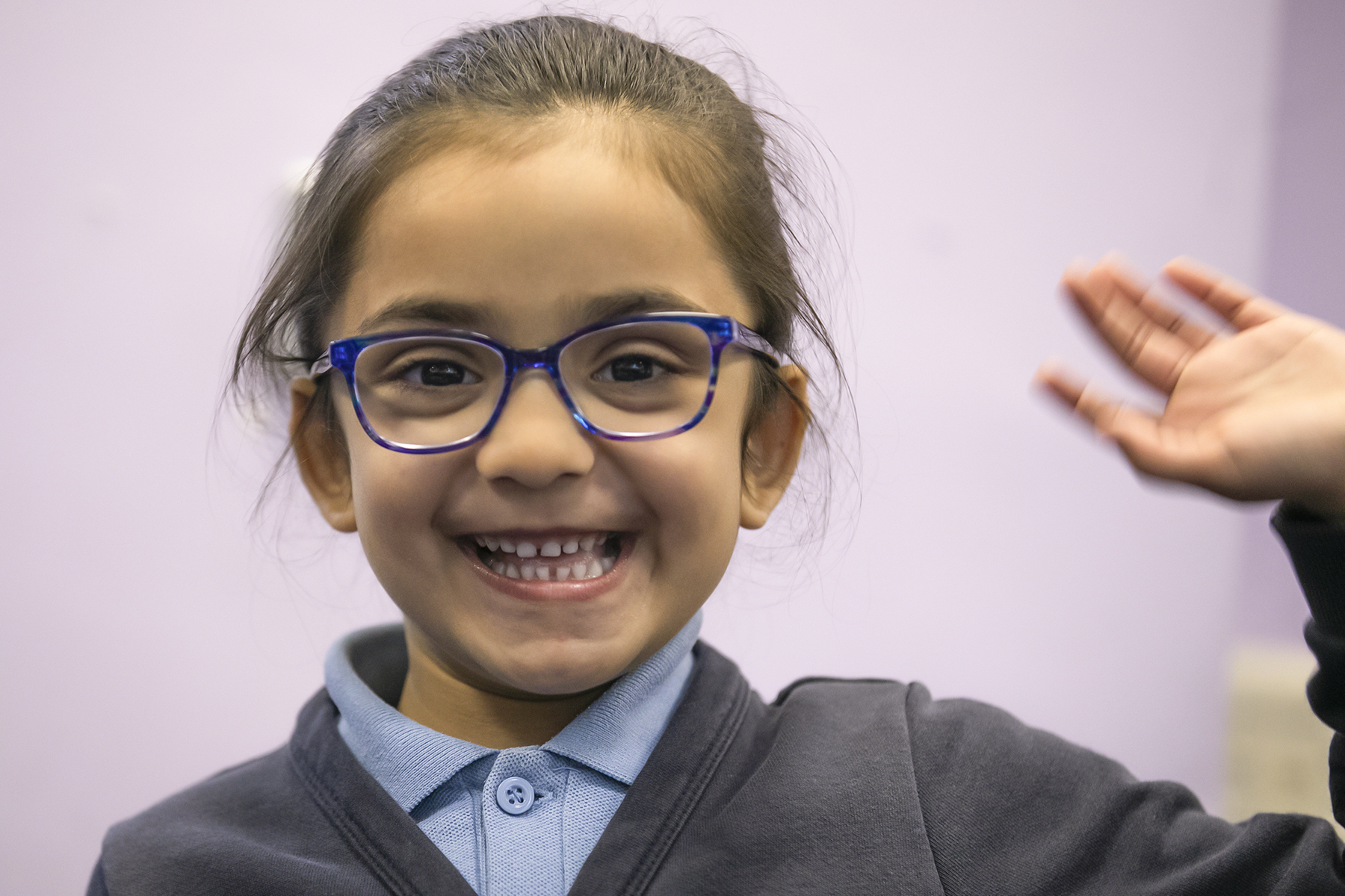 In2Music.GM_.Singlegate.23.COL_.sm_ A young girl with blue glasses smiles widely and waves her hand, excited after attending a CPD workshop. She is wearing a navy blue sweater over a collared shirt, set against a light purple background.