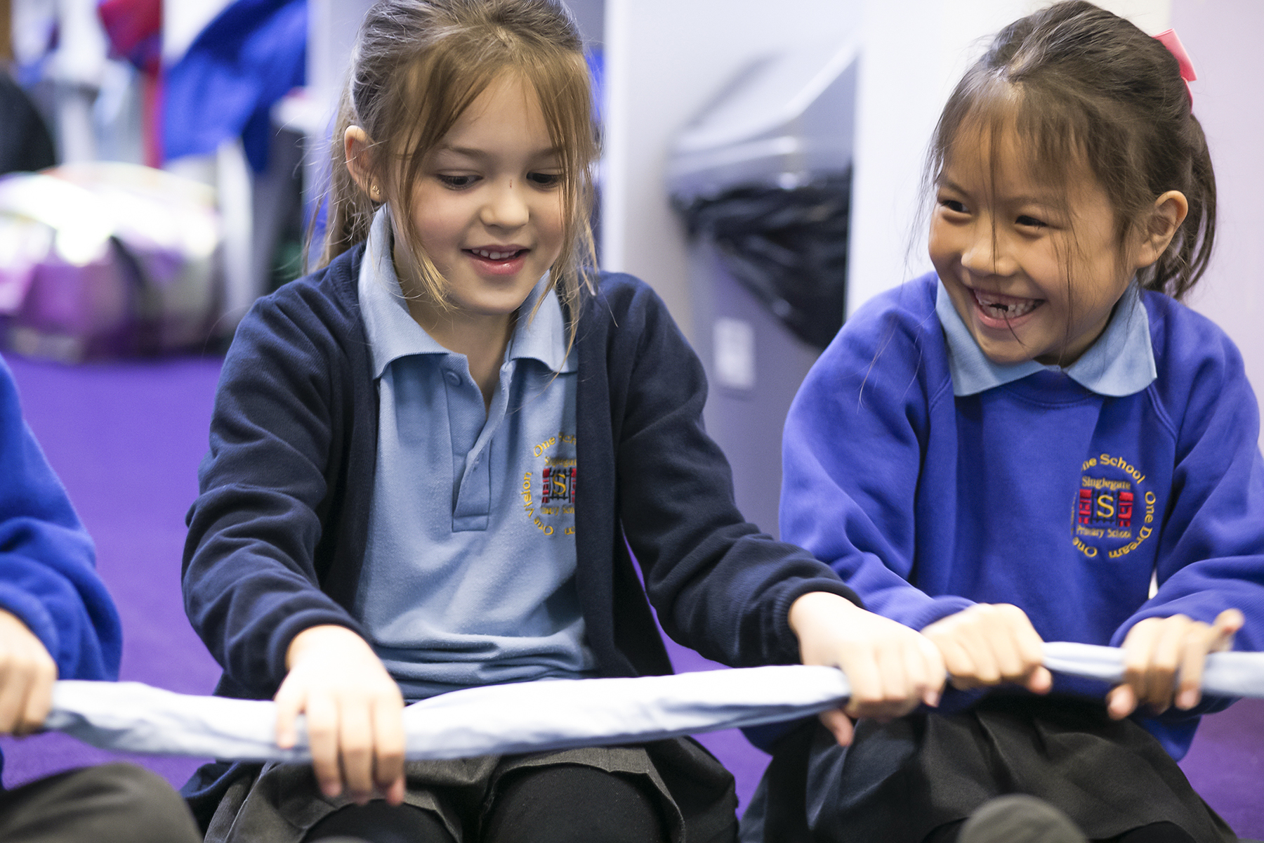 In2Music.GM_.Singlegate.30.COL_.sm_ Two young girls in blue school uniforms smile and laugh while sitting together, holding a piece of fabric in a classroom during a CPD activity.