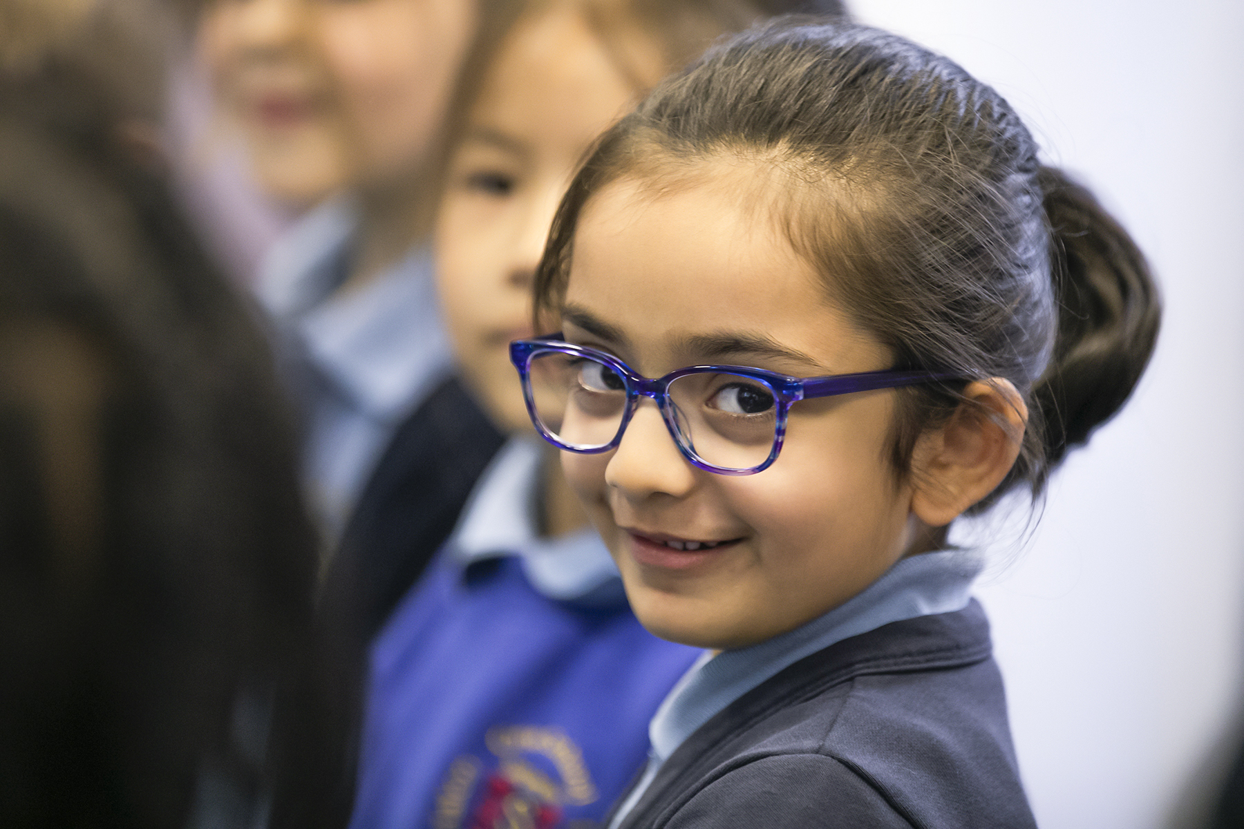 In2Music.GM_.Singlegate.35.COL_.sm_ A young girl with dark hair tied back and wearing blue glasses smiles at the camera while standing among other children in school uniforms during a CPD activity.
