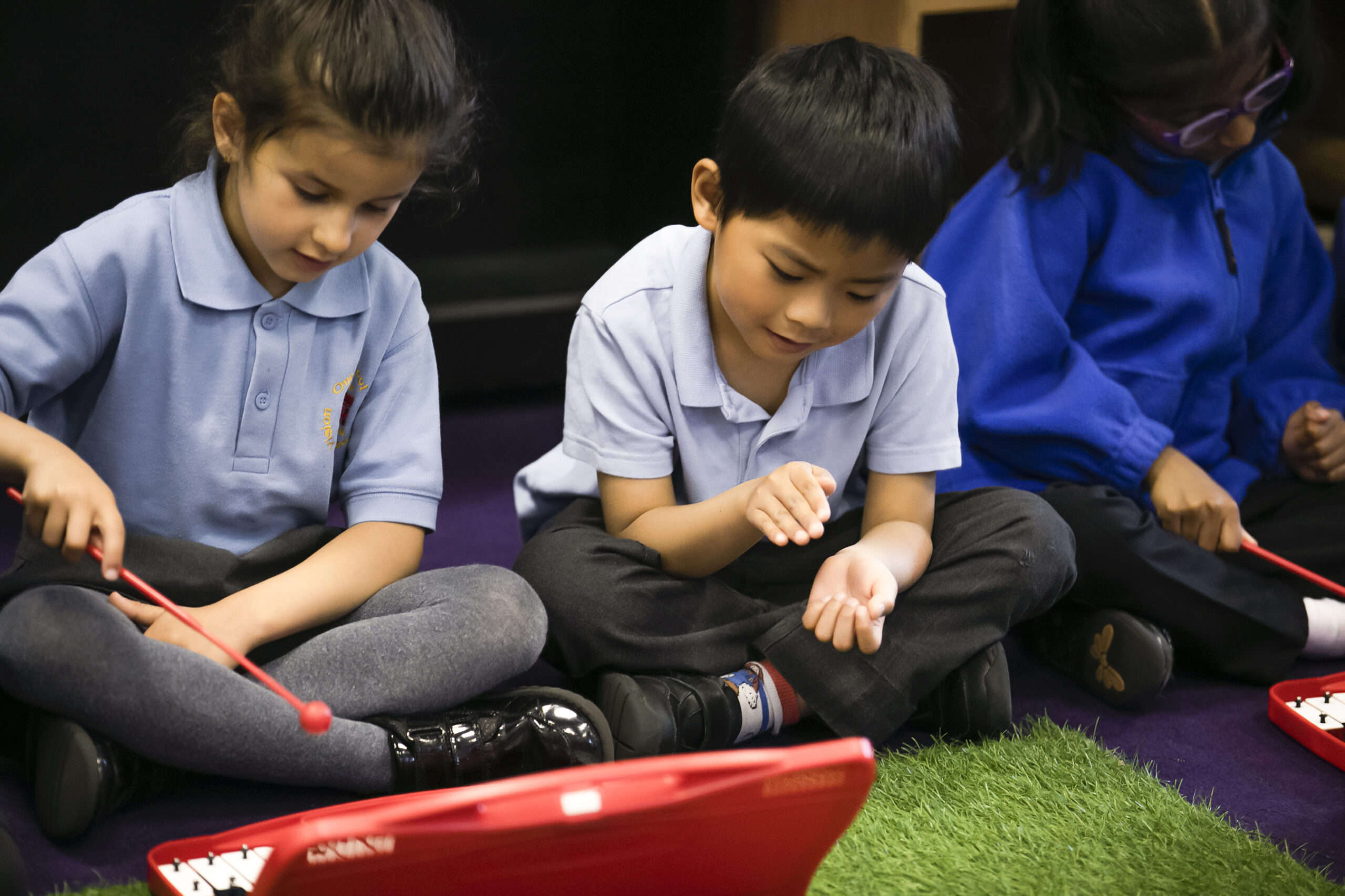 In2Music.GM_.Singlegate.46.COL_.sm_ Three young children sit cross-legged on the floor, playing red percussion instruments with sticks as part of a CPD-inspired music activity. They are focused, in school uniforms, within a classroom featuring a purple carpet and green turf.