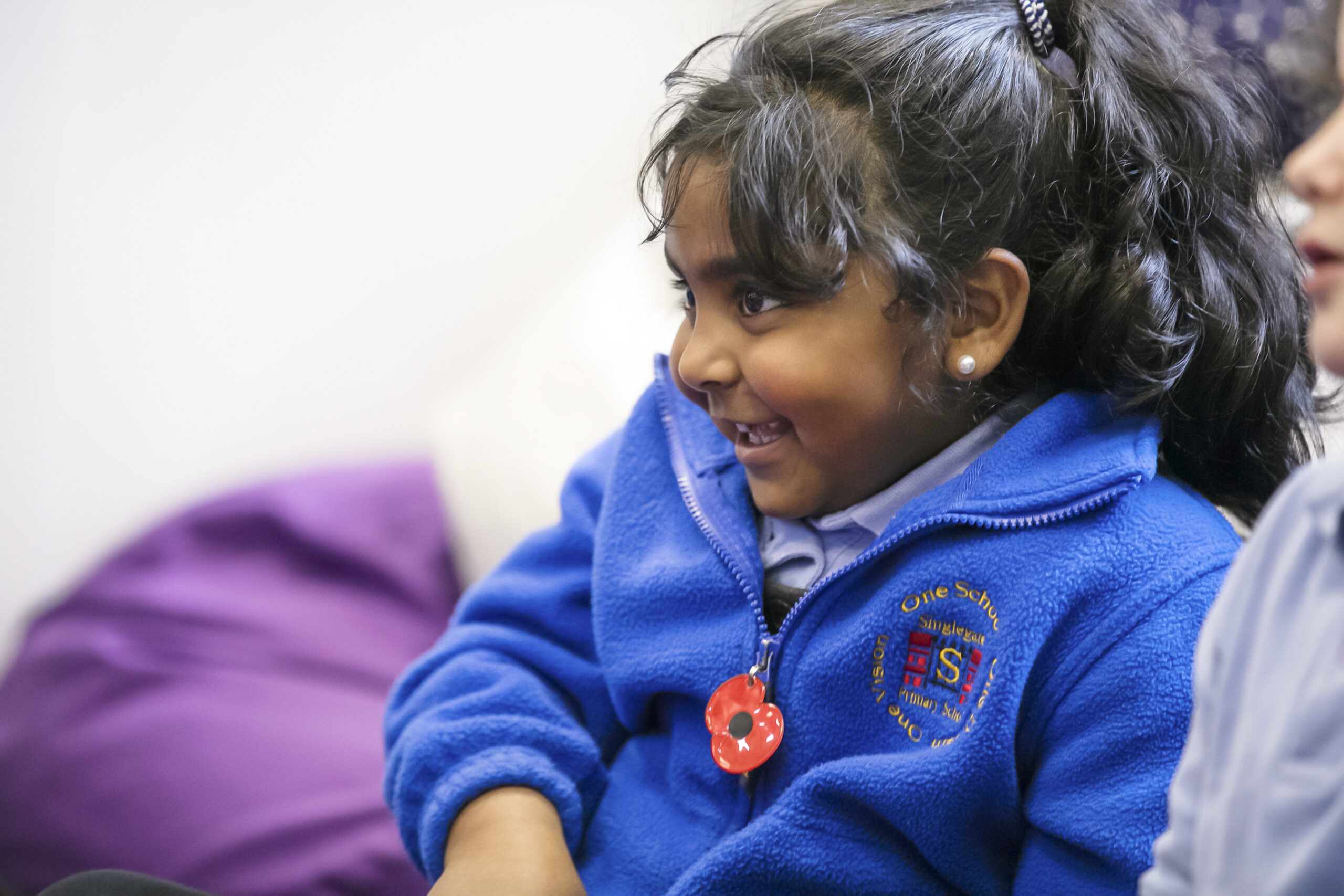 In2Music.GM_.Singlegate.57.COL_.sm_ A young girl with dark hair in a ponytail smiles while wearing a bright blue fleece with a school emblem and a red poppy pin, possibly at a CPD event. She is sitting indoors, looking towards someone off camera.