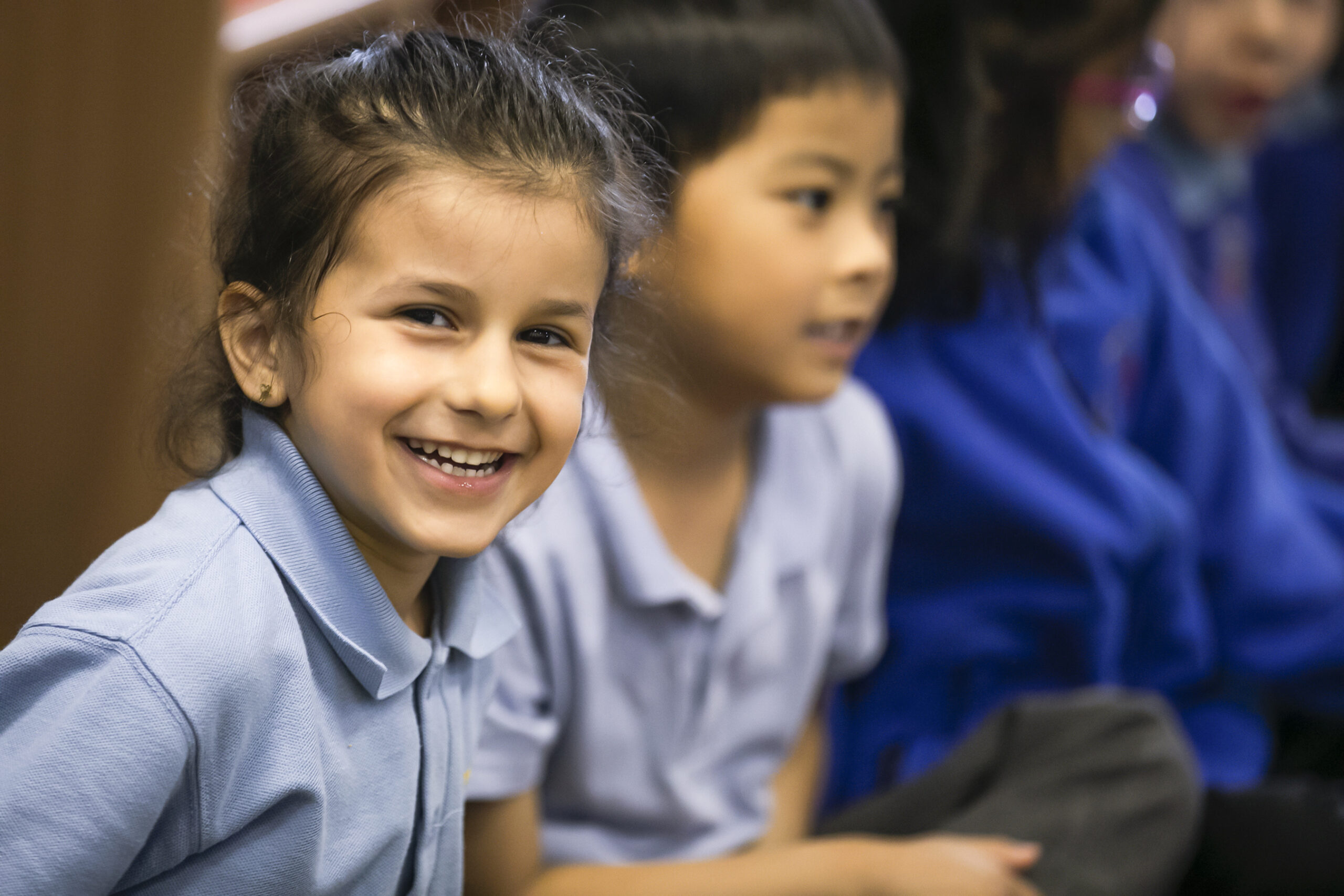 In2Music.GM_.Singlegate.58.COL_.sm_ A young girl in a light blue shirt smiles brightly at the camera while sitting next to other children during a CPD classroom session. The background shows other kids slightly out of focus.