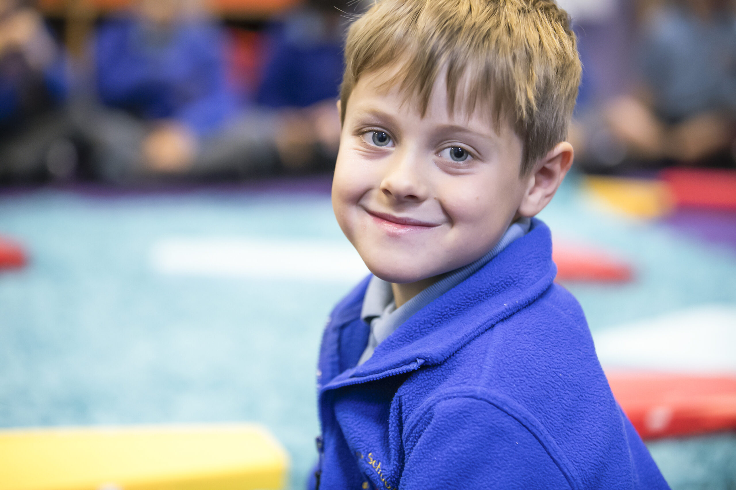 In2Music.GM_.Singlegate.64.COL_.sm_ Young boy with light brown hair wearing a bright blue fleece smiles at the camera while sitting in a CPD classroom setting with colorful mats and blurred classmates in the background.