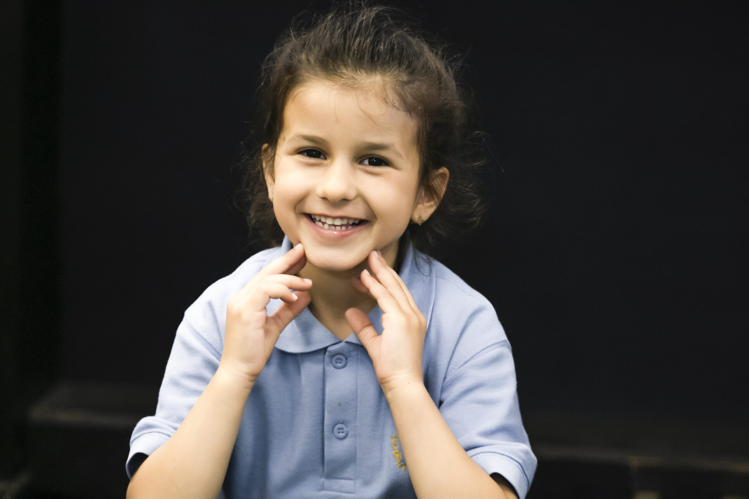 In2Music.GM_.Singlegate.65.COL_.sm_ A young girl with dark hair smiles brightly, holding her hands near her chin. She is wearing a light blue polo shirt, perhaps attending a CPD event, and sits against a dark background.