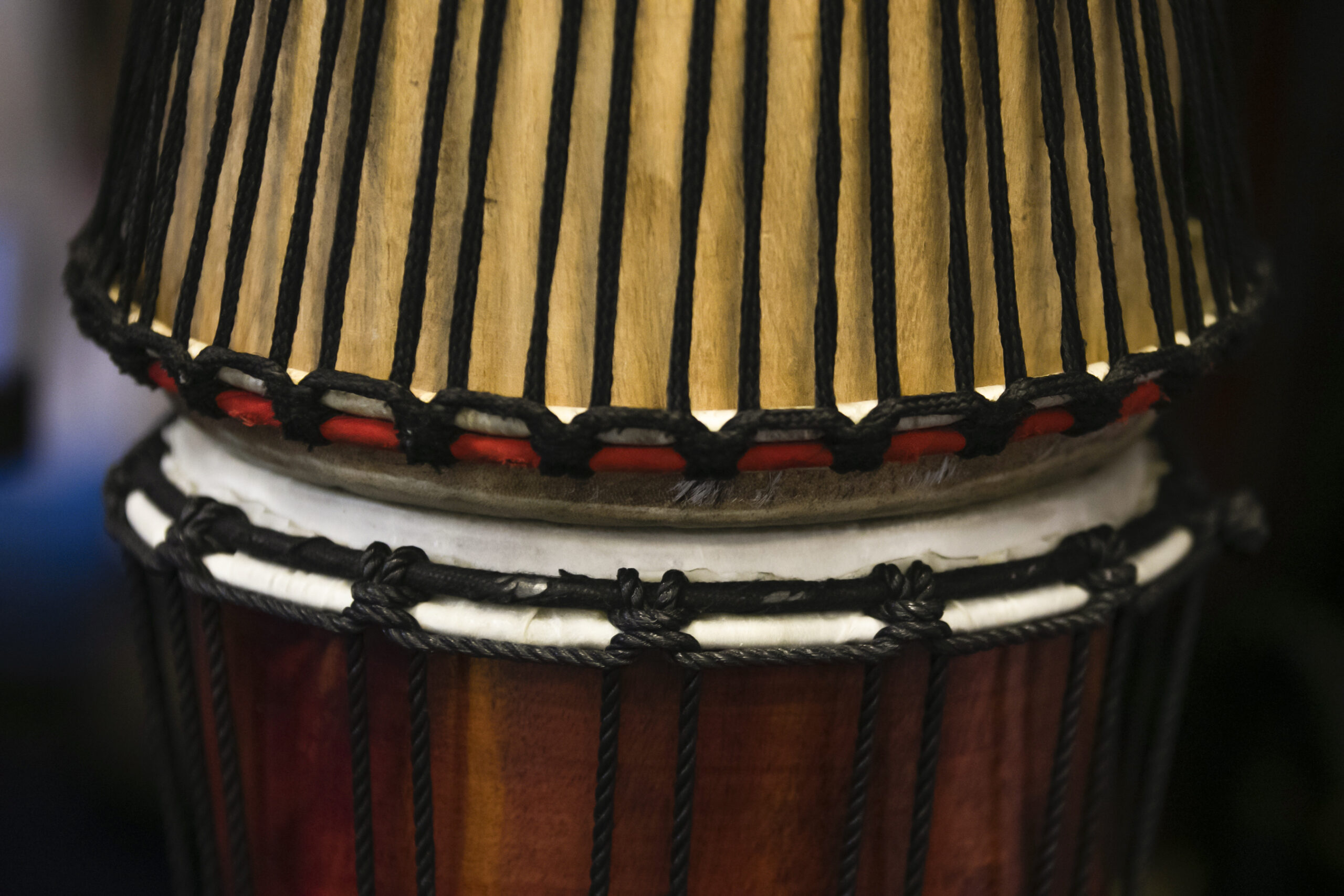 In2Music.GM_.Singlegate.68.COL_.sm_ Close-up of the top section of a traditional African drum, highlighting its wooden body, tightly woven black ropes, CPD red accents, and white drumhead.