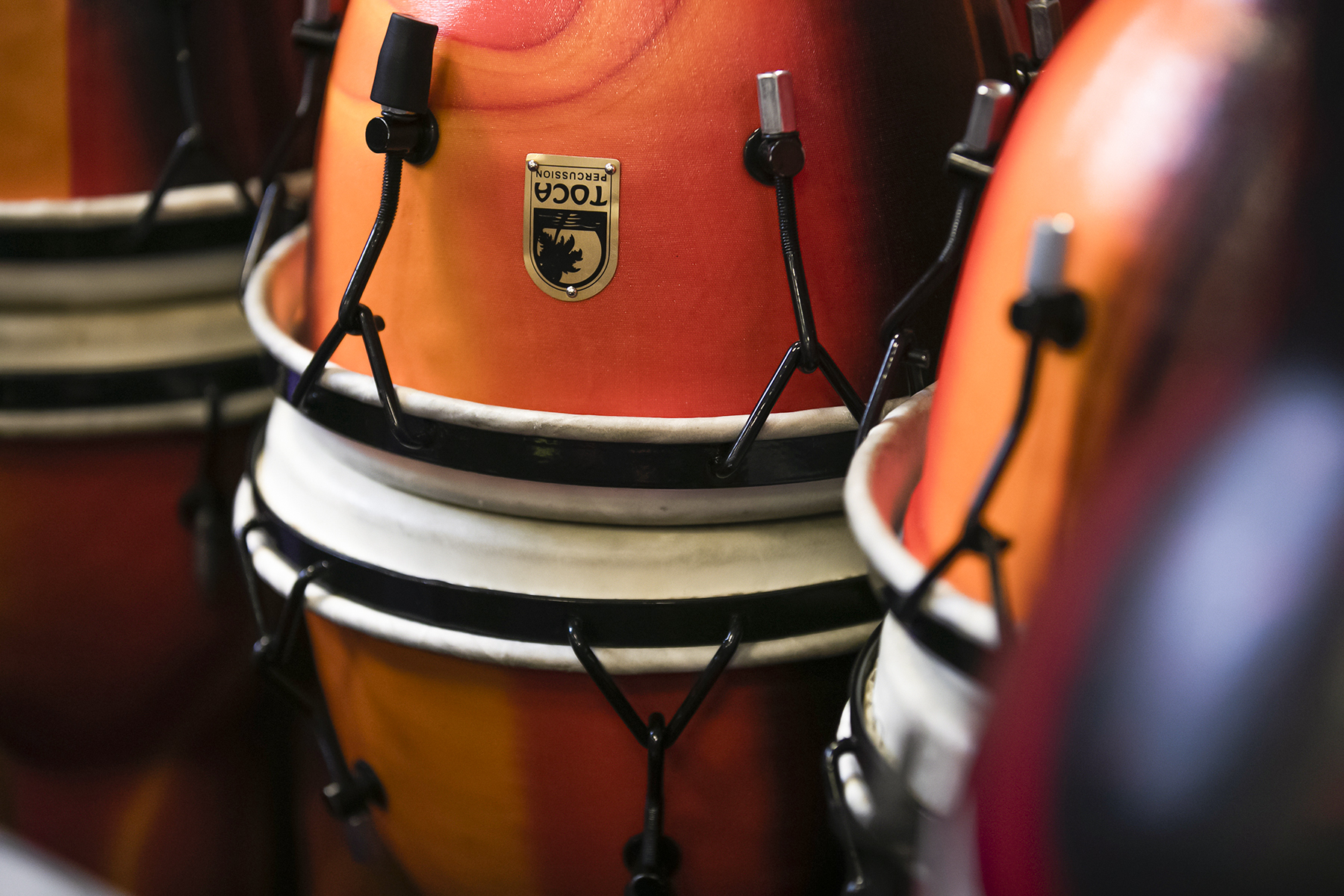 In2Music.GM_.Singlegate.69.COL_.sm_ Close-up of several orange and red Toca brand CPD conga drums, showing their glossy finish, metal hardware, and white drumheads. The drums are tightly arranged, emphasizing their vibrant colors and textures.