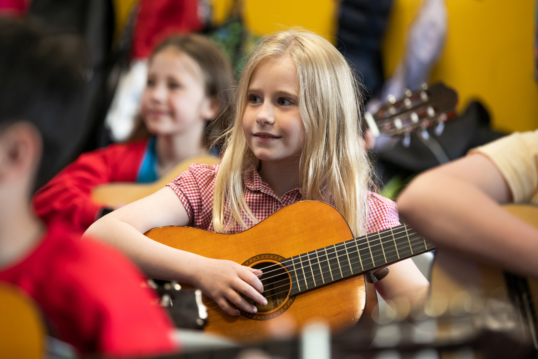 In2Music.Guitars.JosephHood.02.sm_ A young girl with long blonde hair smiles while playing an acoustic guitar in a classroom CPD session, surrounded by other children also holding guitars.