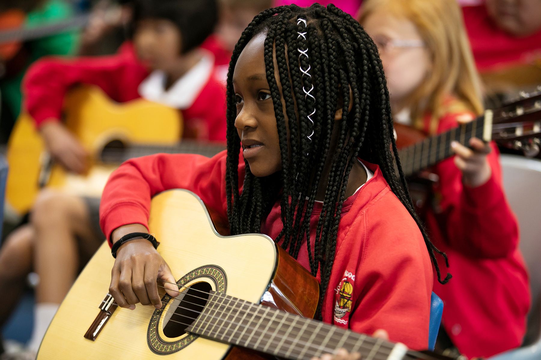In2Music.Guitars.JosephHood.05.sm_ A young girl with braided hair, wearing a red school uniform, sits and plays an acoustic guitar in a classroom during a CPD music session, with other children also playing guitars in the background.