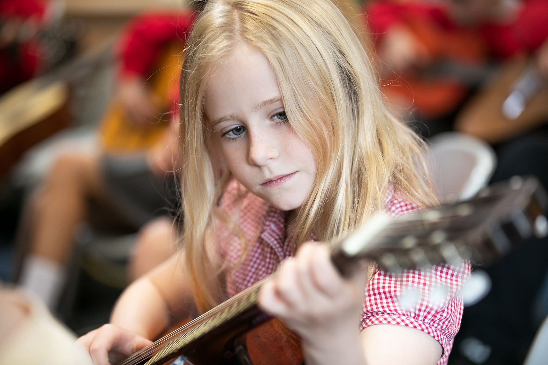 In2Music.Guitars.JosephHood.08.sm_ A young girl with long blonde hair, wearing a red and white checkered shirt, focuses intently as she plays an acoustic guitar during a CPD music session. Other children playing instruments are blurred in the background.