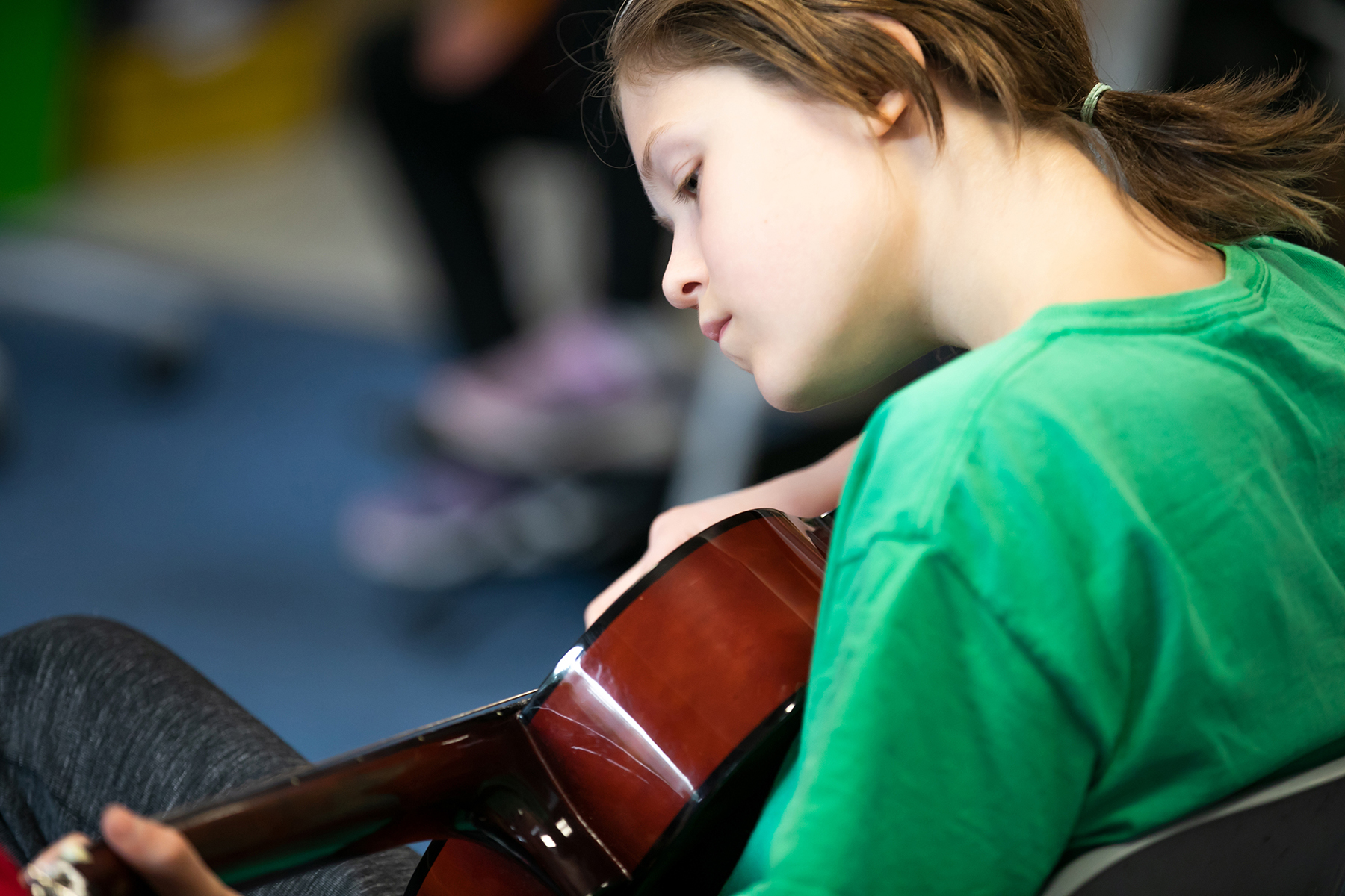 In2Music.Guitars.JosephHood.10.sm_ A young person in a green shirt sits and plays an acoustic guitar, concentrating on the instrument. The background is blurred, drawing focus to their thoughtful expression and the guitar—a moment of personal CPD through music.