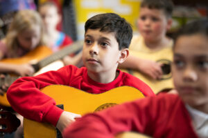 In2Music.Guitars.JosephHood.13.sm_ A young boy in a red sweatshirt plays an acoustic guitar in a classroom during a CPD session, surrounded by other children holding guitars, all attentively watching something out of frame.