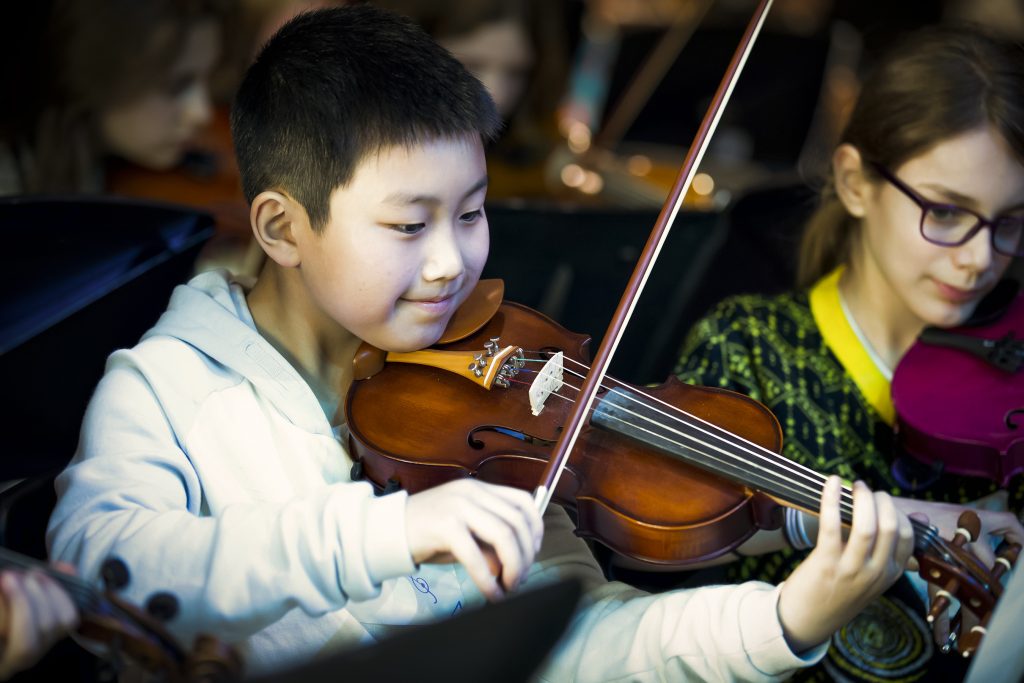MMF.Magic_.120.COL_-scaled A young boy in a white hoodie plays the violin, smiling and focused, while a girl with glasses beside him also plays. Donate to help more children enjoy music; other kids and instruments appear blurred in the background.