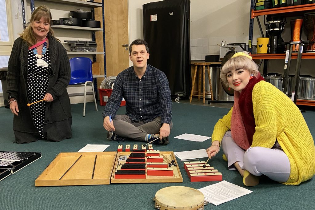 MMF_CPD Three adults sit on the floor in a music room, smiling at the camera during a CPD session. They are surrounded by xylophones, percussion instruments, and scattered sheet music. Shelves of drums and other instruments are visible behind them.