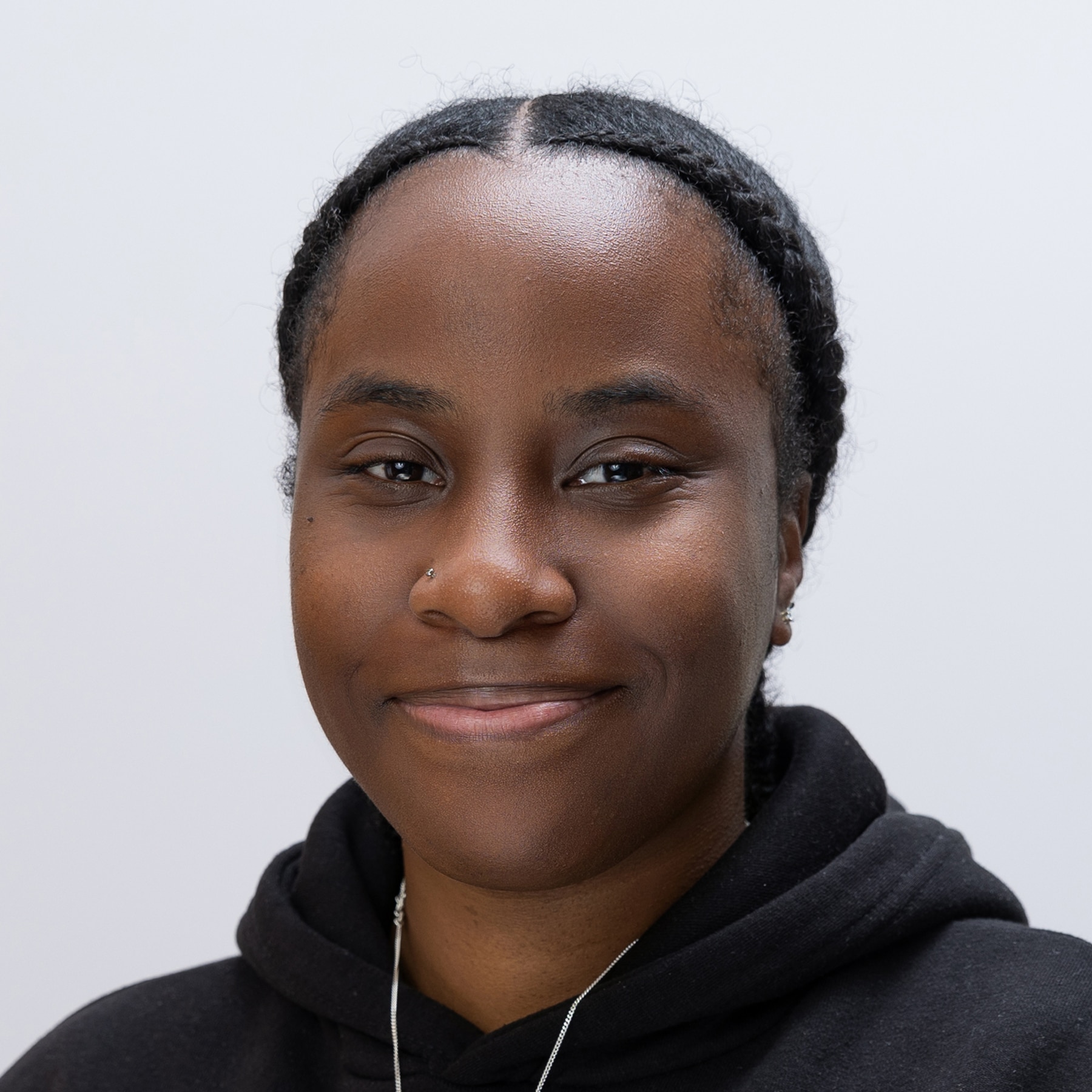 Meet the team: A woman with braided hair, wearing a black hoodie and a necklace, smiles gently against a plain light background.