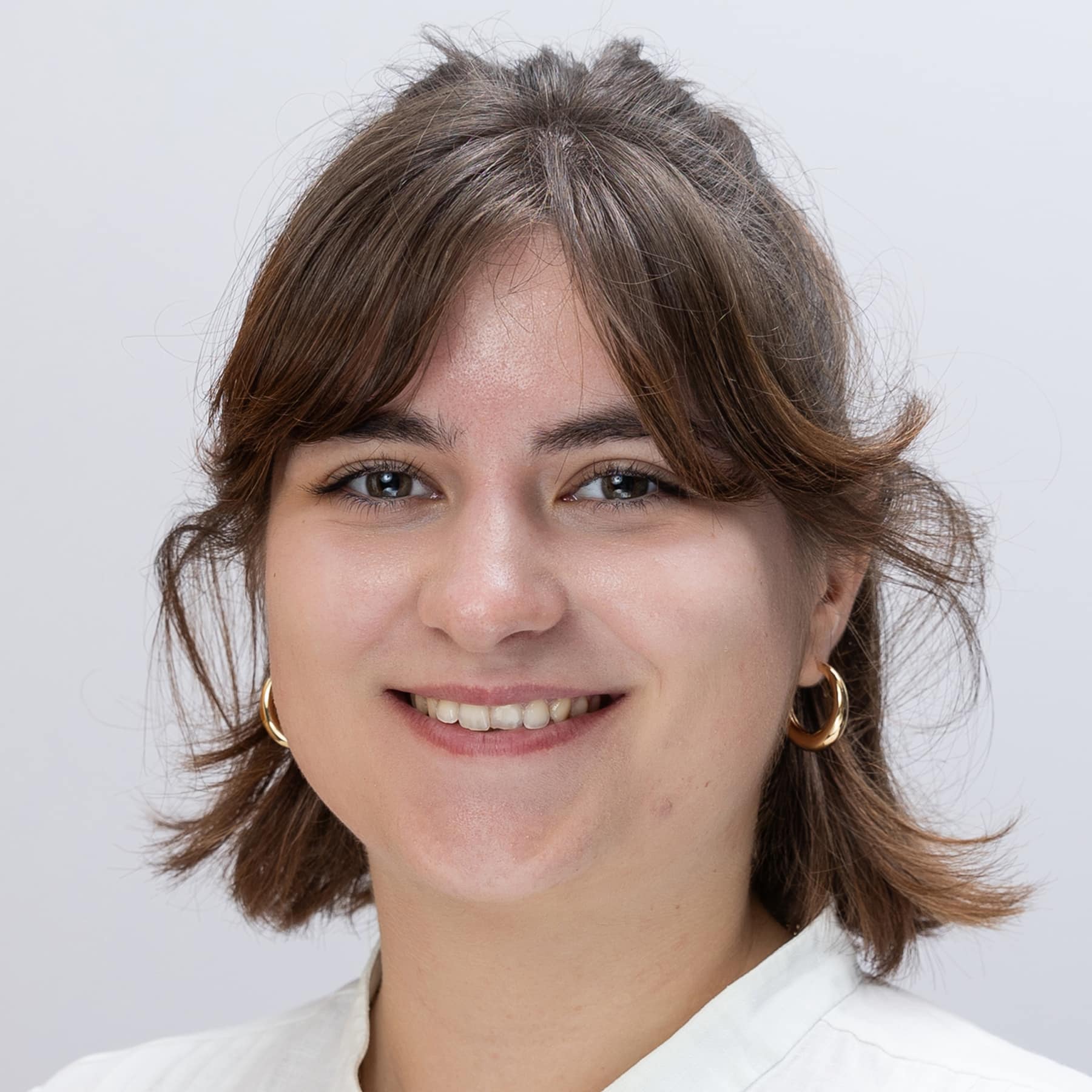 Meet the team: A young woman with short brown hair and bangs smiles at the camera. She is wearing gold hoop earrings and a white top, set against a light gray background.