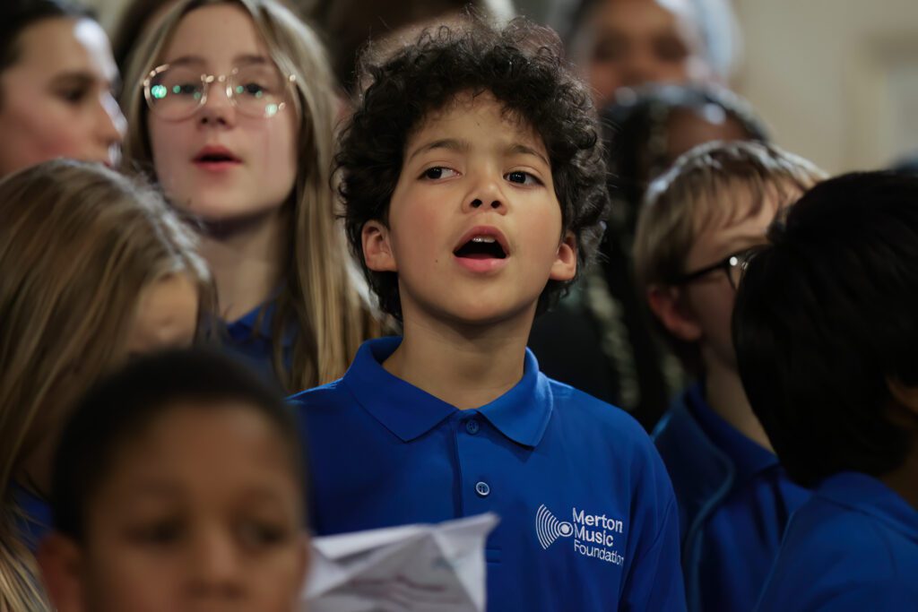 A young boy in a blue Merton Music Foundation shirt sings with a group of children in choirs, some wearing glasses, during a choir performance. The background is softly blurred, focusing on the children's faces.