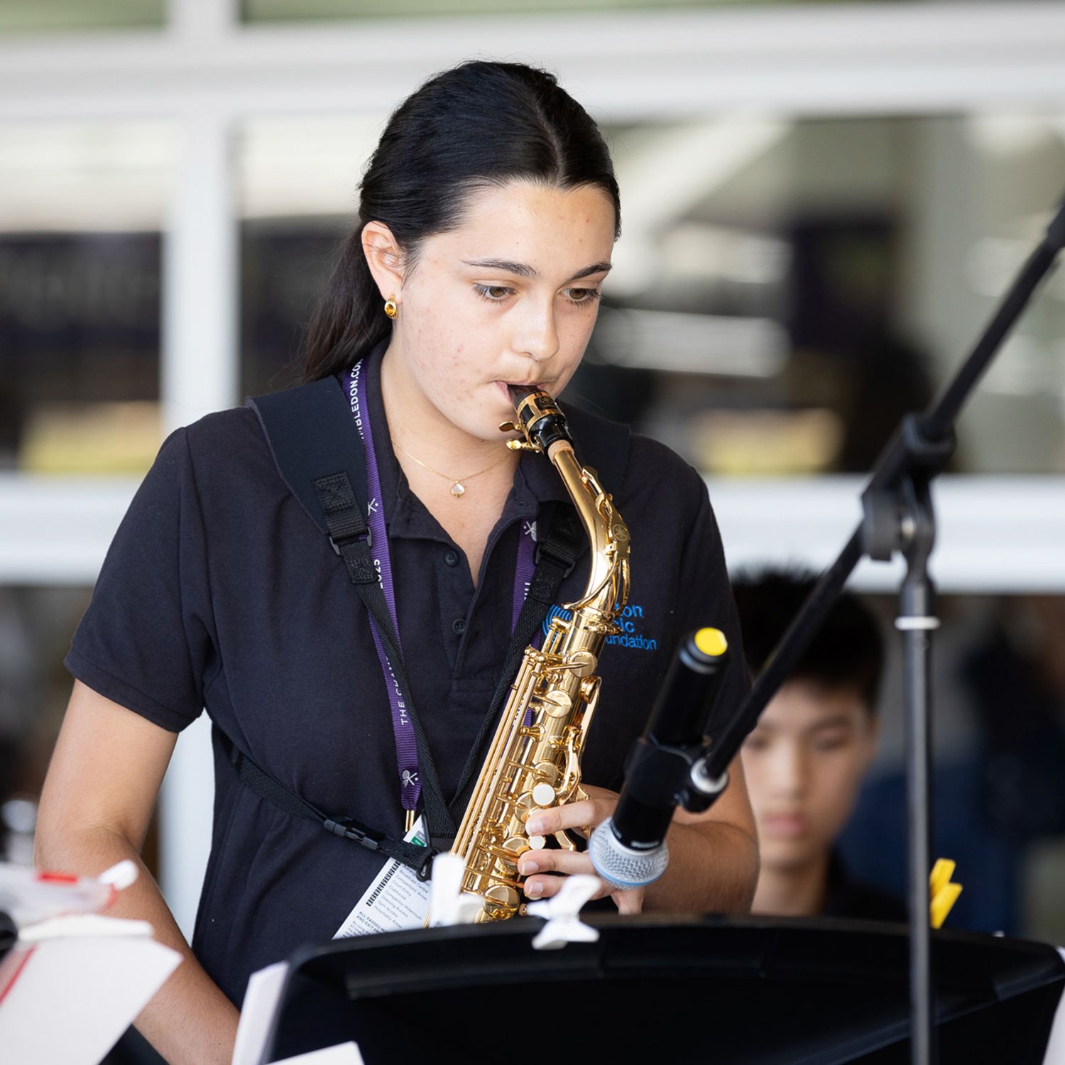 A young woman wearing a black polo shirt plays an alto saxophone in front of a microphone, with sheet music in front of her and another person blurred in the background.