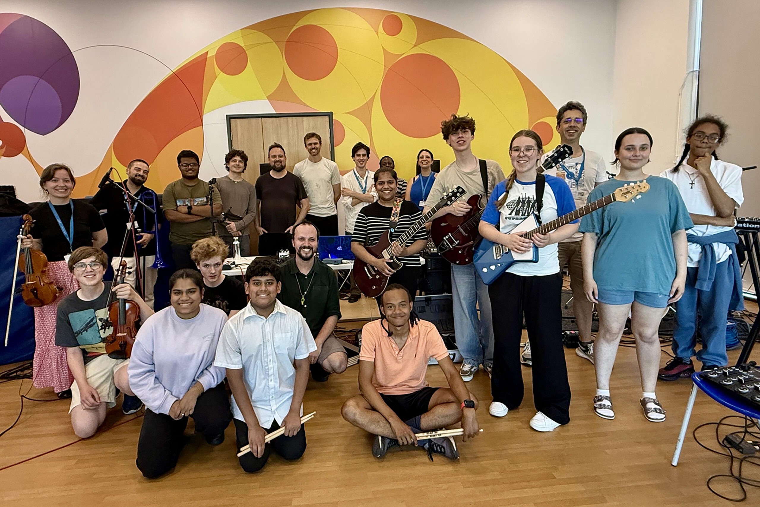 A diverse group of young people and adults pose together in a bright room with colorful wall art. Some hold musical instruments like guitars and violins. Everyone is smiling, showing a sense of camaraderie and excitement.