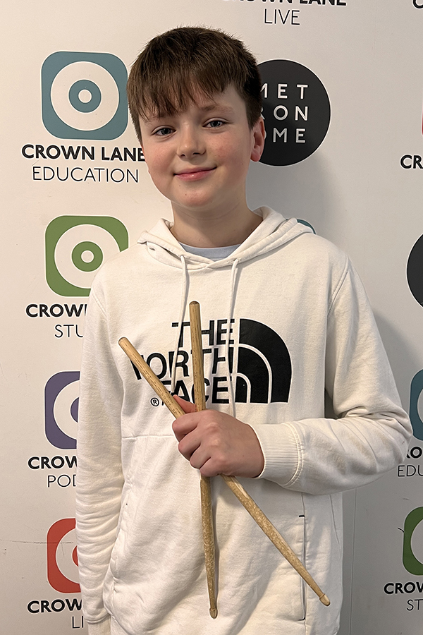 A boy in a white hoodie holds two drumsticks and smiles at the camera. He stands in front of a wall with Crown Lane Education and Metronome Live logos, making this shot a standout addition to music-themed photo collections.