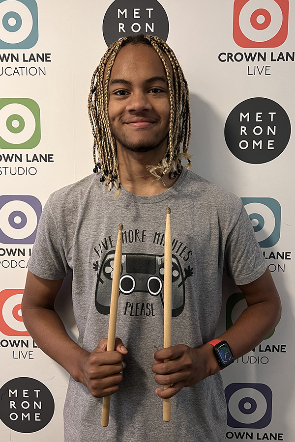 A smiling person with blonde plaits holds drumsticks and stands in front of a wall with Metronome, Crown Lane Live, and Crown Lane Studio logos—captured as part of our vibrant Photo Collections. They wear a grey t-shirt and a red smartwatch.