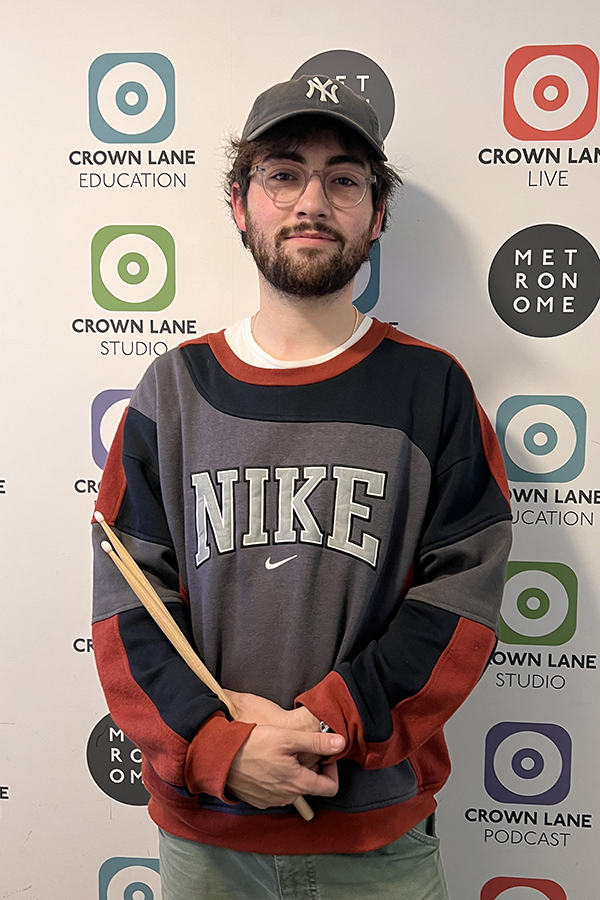 A young man wearing a Nike sweatshirt and a NY Yankees cap holds drumsticks and stands in front of a wall with Crown Lane Studio, Education, Live, Metronome Podcast logos—a standout shot for music-themed photo collections.