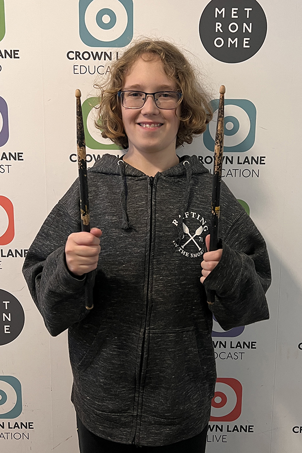 A smiling child with glasses and shoulder-length hair holds two drumsticks upright. They wear a dark zip-up hoodie, standing before a wall with colourful Crown Lane and Metronome logos—perfect for vibrant Photo Collections.