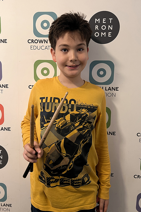 A smiling boy with short dark hair, wearing a yellow Turbo shirt with a car design, holds two drumsticks. He stands in front of a wall with Crown Lane Education and Metronome logos—perfect for Photo Collections.