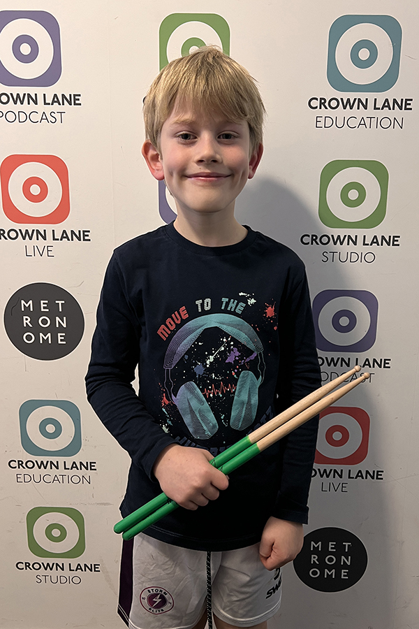 A young boy with blonde hair and a dark shirt holds drumsticks and smiles in front of a backdrop featuring Crown Lane Education, Studio, Podcast, and Live logos in circles—a perfect addition to Photo Collections.