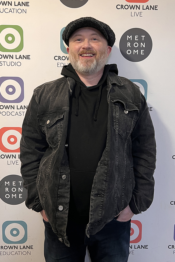 A smiling man with a short light beard, wearing a black flat cap, hoodie, and denim jacket, stands in front of a wall featuring circular logos like Crown Lane and Metronome—a great addition to your Photo Collections.