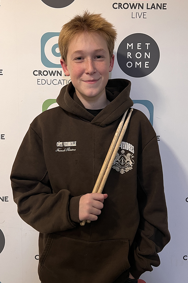A smiling boy with short, light brown hair holds two drumsticks and wears a dark hoodie with logos. He stands before a backdrop showing Crown Lane Education and Metronome logos, perfect for engaging Photo Collections.
