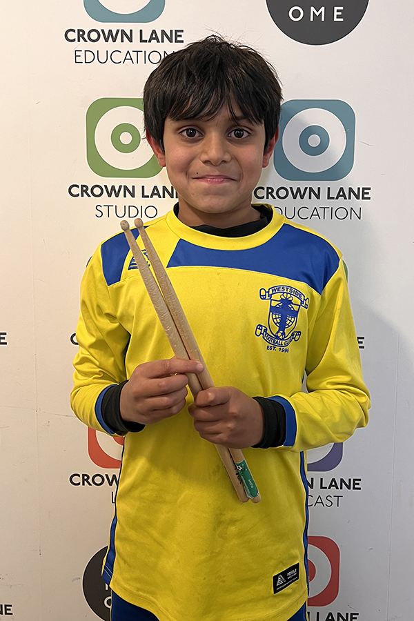 A young boy in a yellow sports jersey holds two drumsticks and smiles, standing in front of a wall with multiple Crown Lane logos—perfect for music-themed Photo Collections.