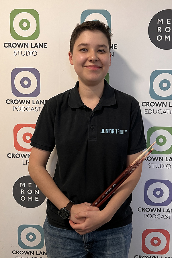 A young person with short hair smiles whilst holding drumsticks. Wearing a black Junior Trinity shirt, they stand by a backdrop with logos for Crown Lane Studio, Podcast, Education, and Metronome—perfect for adding to Photo Collections.