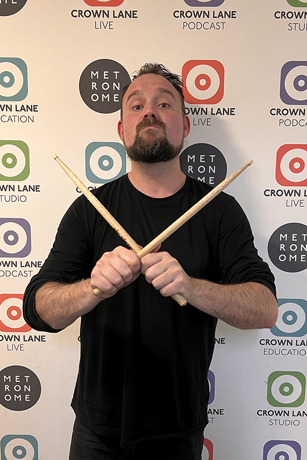 A bearded man in a black shirt holds two drumsticks crossed in front of his chest, standing against a backdrop with Crown Lane and Metronome logos—perfect for music-themed photo collections.