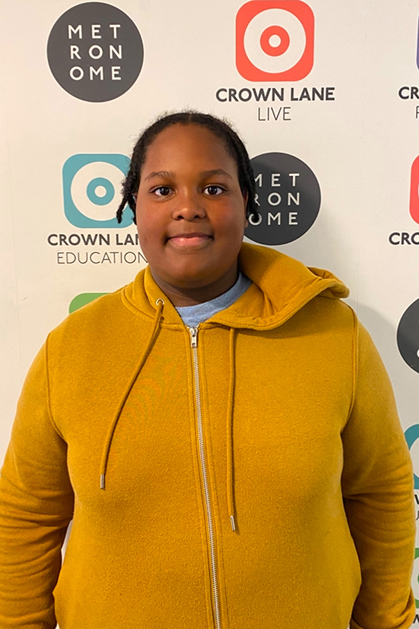 A person wearing a mustard yellow zip-up hoodie stands smiling in front of a wall with Metronome and Crown Lane logos, perfect for Photo Collections capturing vibrant moments.