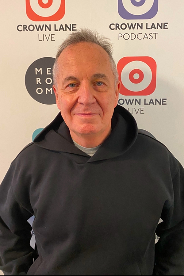 A man in a black hoodie stands in front of a white backdrop featuring Crown Lane Live, Crown Lane Podcast, and Me Room logos, smiling slightly at the camera—a great addition to your photo collections.