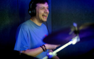A smiling man wearing headphones and a blue shirt plays drums in a dimly lit room with blue lighting.