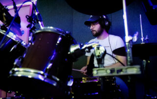 A man wearing glasses, a cap, and headphones plays a drum kit in a music studio, surrounded by drums and cymbals, focused on his performance.