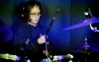 A person wearing glasses and headphones plays a drum kit, holding drumsticks and concentrating, with blue and green lighting in the background.