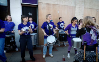 A group of women, mostly wearing purple shirts, play drums together on a pavement in an urban area. One woman faces the group and leads them. Wooden panels cover part of the building behind them.