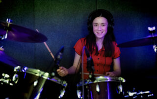 A smiling girl wearing headphones and a red shirt plays a drum kit in a soundproof room, with drumsticks in her hands and cymbals visible in the foreground.