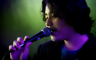 A person with medium-length dark hair is singing into a microphone, eyes closed, under purple and green stage lighting.
