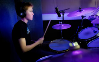 A young person wearing headphones plays a drum kit in a music studio with purple lighting, focused on the drums with a microphone positioned above the cymbals.