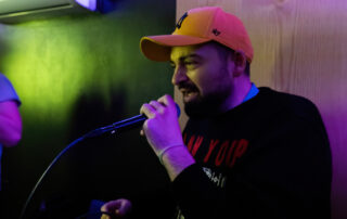 A man wearing a yellow cap and black jumper sings into a microphone in a dimly lit room, with colourful lighting casting purple and green hues on the scene.
