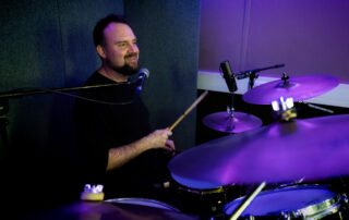 A man with a beard plays a drum kit in a recording studio, smiling as he holds drumsticks. There are microphones set up around the drums, and the lighting is purple and blue.