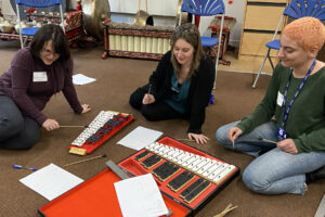 IMG_3171.cr Three people sit on a carpeted floor, playing xylophones with beaters. Sheet music and papers are scattered nearby, suggesting a collaborative music activity often found in schools.