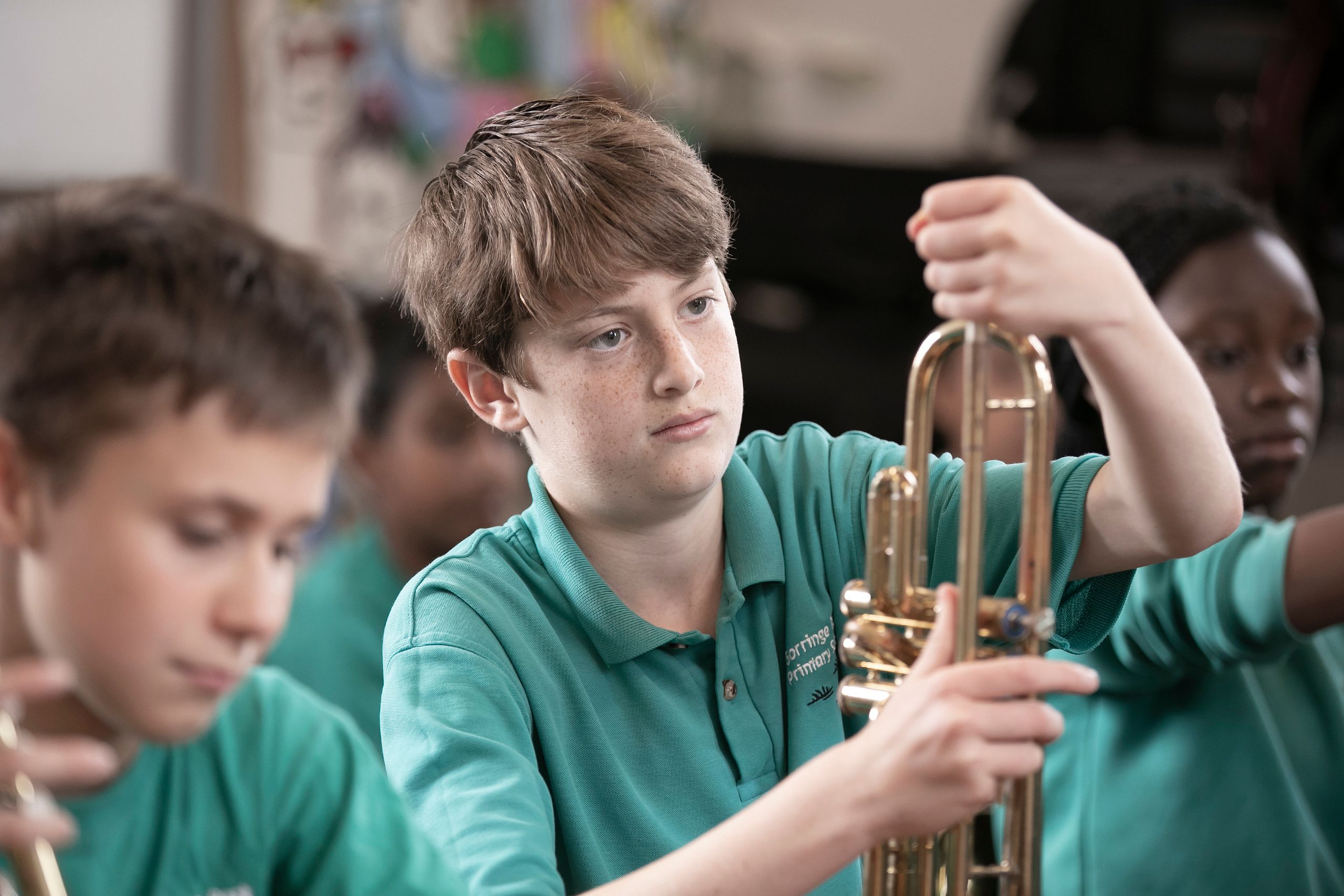 A boy in a teal shirt is assembling or tuning a brass instrument, likely a trumpet, in a classroom setting with other students around him, also wearing teal shirts.
