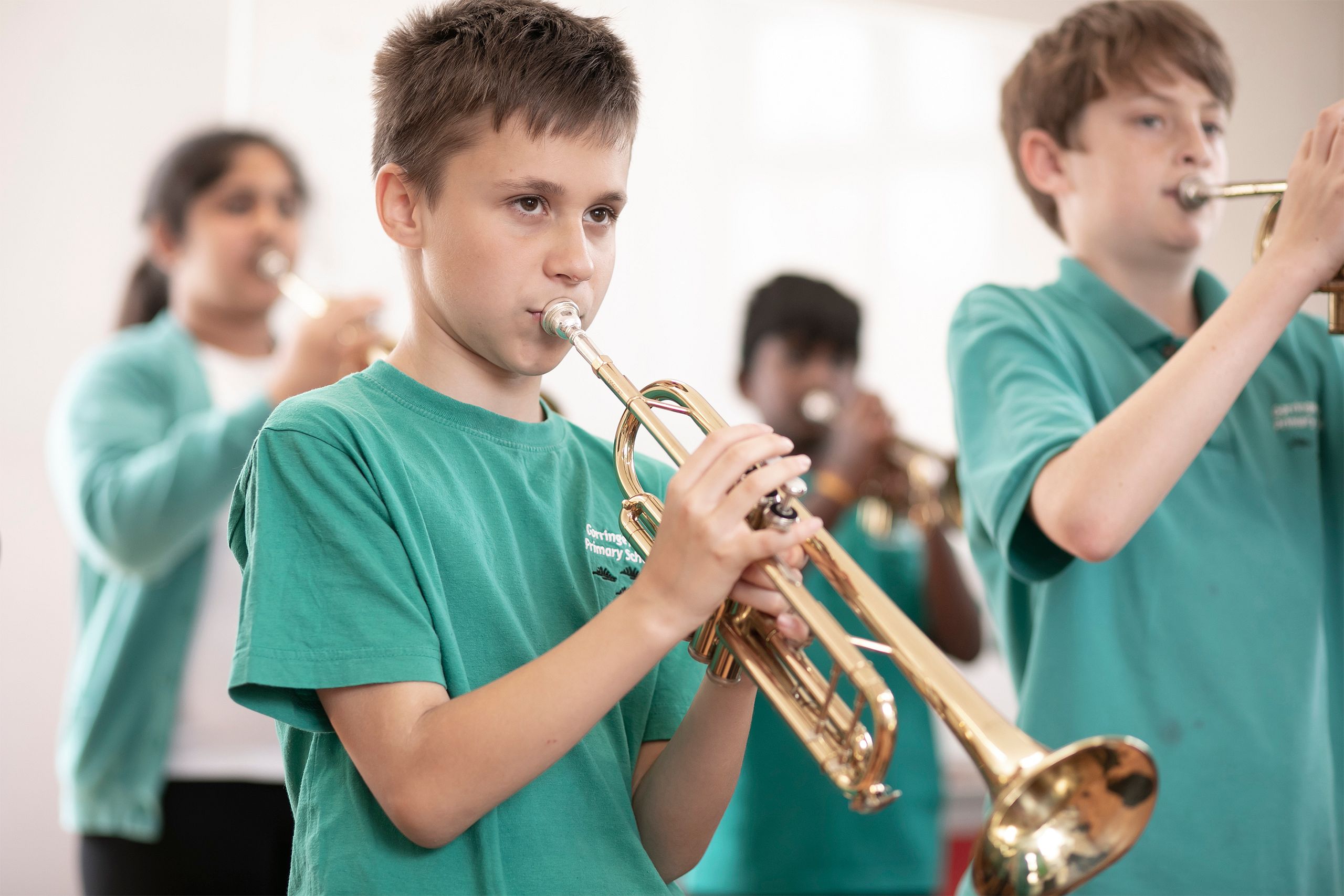 A group of children in teal shirts play trumpets indoors, focusing intently. The background is softly blurred, emphasising the children and their instruments in the foreground.